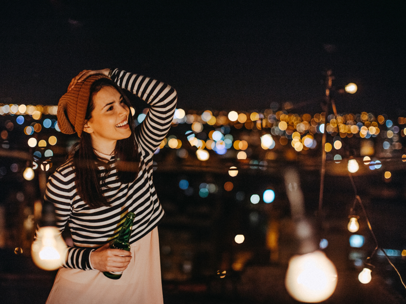 Eine Frau steht mit einer Flasche in der Hand auf einer Dachterrasse während einer Party.