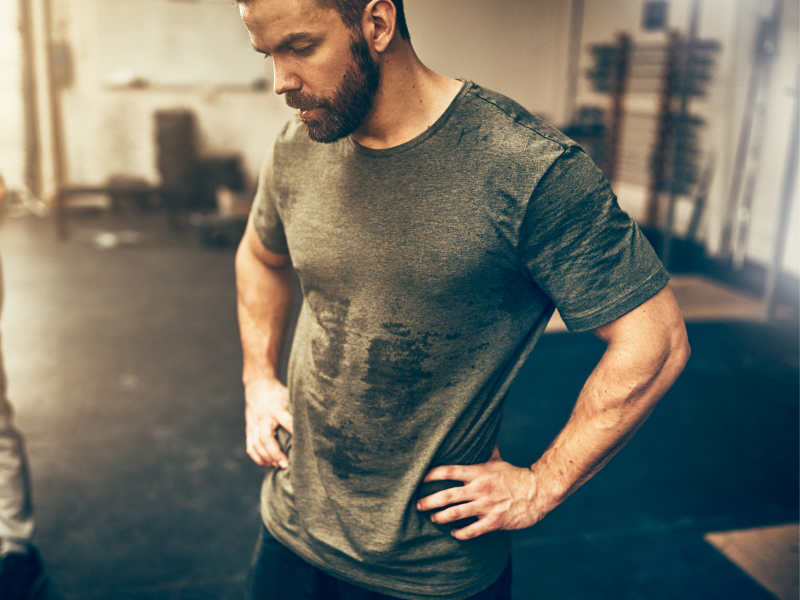 Mann mit Schweißflecken auf dem Shirt steht in einem Fitnessstudio.
