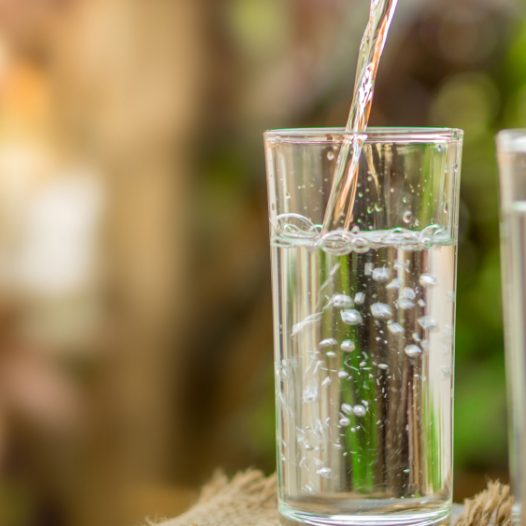 Fokus auf Wasser, das in ein Trinkglas eingegossen wird. Ein volles Glas steht daneben. Natur verschwommen im Hintergrund.