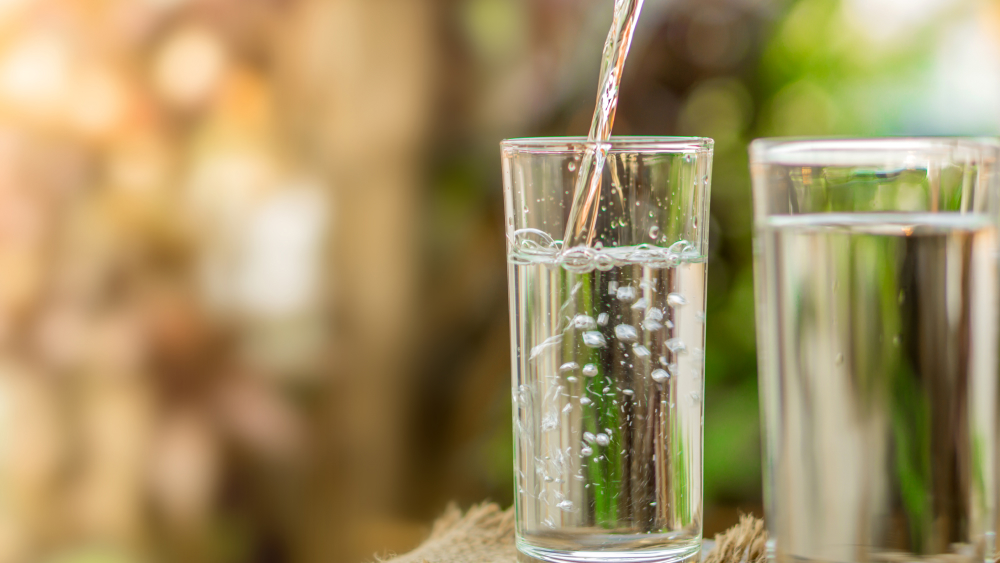 Fokus auf Wasser, das in ein Trinkglas eingegossen wird. Ein volles Glas steht daneben. Natur verschwommen im Hintergrund.