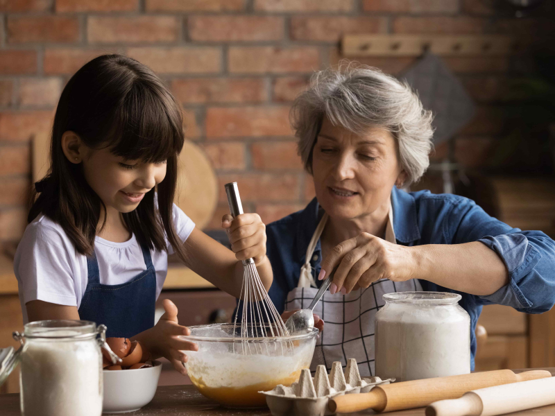 Enkelin und Oma backen zusammen. Die Enkelin rührt den Teig mit einem Schneebesen. Die Oma ergänzt eine Zutat mit einem Löffel.