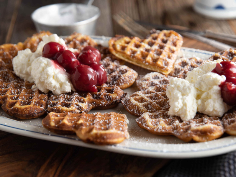 Zwei herzförmige Waffeln mit Puderzucker, Sahne und Sauerkirschen darauf.