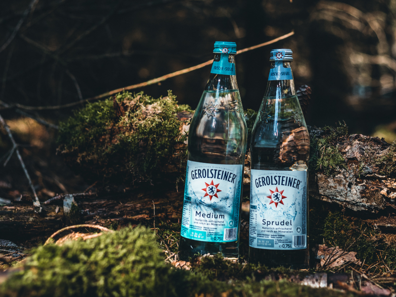Jeweils eine Flasche der Gerolsteiner Mineralwässer Medium und Sprudel stehen im Wald auf moosigem und laubbedecktem Boden.