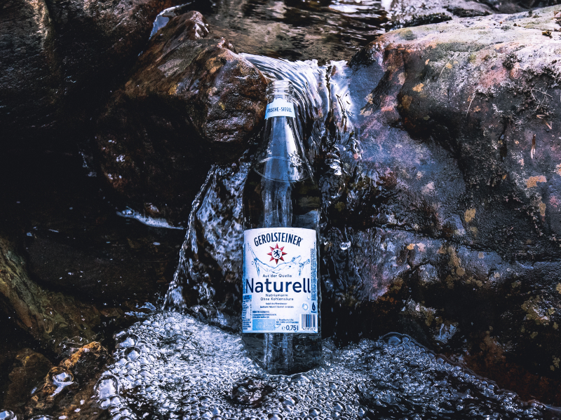 Eine Flasche Gerolsteiner Naturell steht in einer Wasserpfütze, die aufgrund von durch Gestein laufendem Wasser entstanden ist.