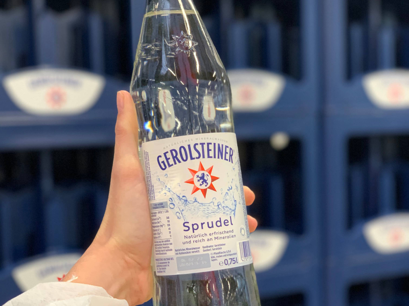 Eine Glasflasche Gerolsteiner Sprudel wird von einer Hand ins Bild gehalten. Im Hintergrund sind blaue Gerolsteiner Wasserkästen zu sehen.