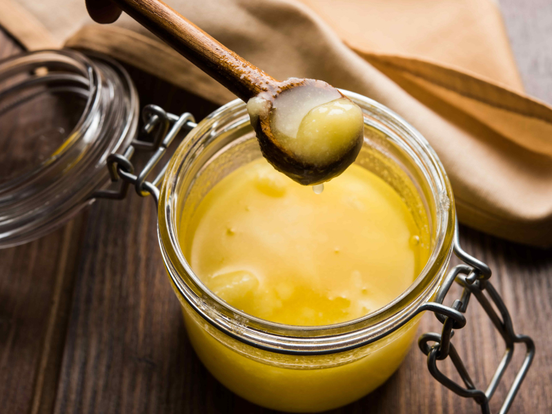 Reines Ghee in einem kleinen Einmachglas mit Deckel. Ein kleiner Holzlöffel mit etwas Ghee wird darüber gehalten.