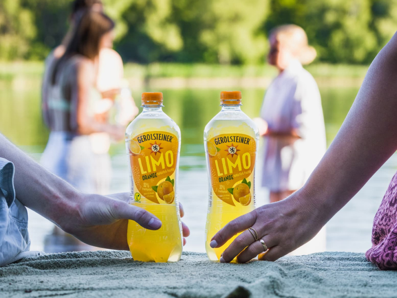Zwei Flaschen Orangenlimonade von Gerolsteiner. Jeweils eine Person greift nach eine der Flaschen. Im Hintergrund sind verschwommen ein See und 3 weitere Personen zu sehen.