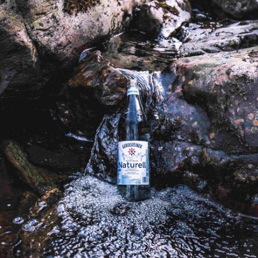 Eine Gerolsteiner Naturell Glasflasche steht auf einem Felsvorsprung eines Bachverlaufs in der Eifel.