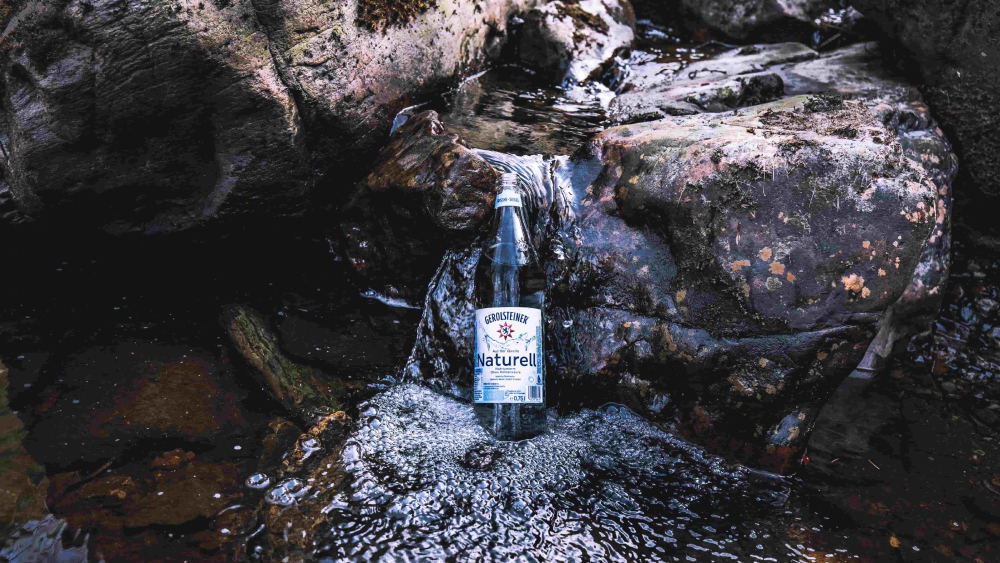 Eine Gerolsteiner Naturell Glasflasche steht auf einem Felsvorsprung eines Bachverlaufs in der Eifel.