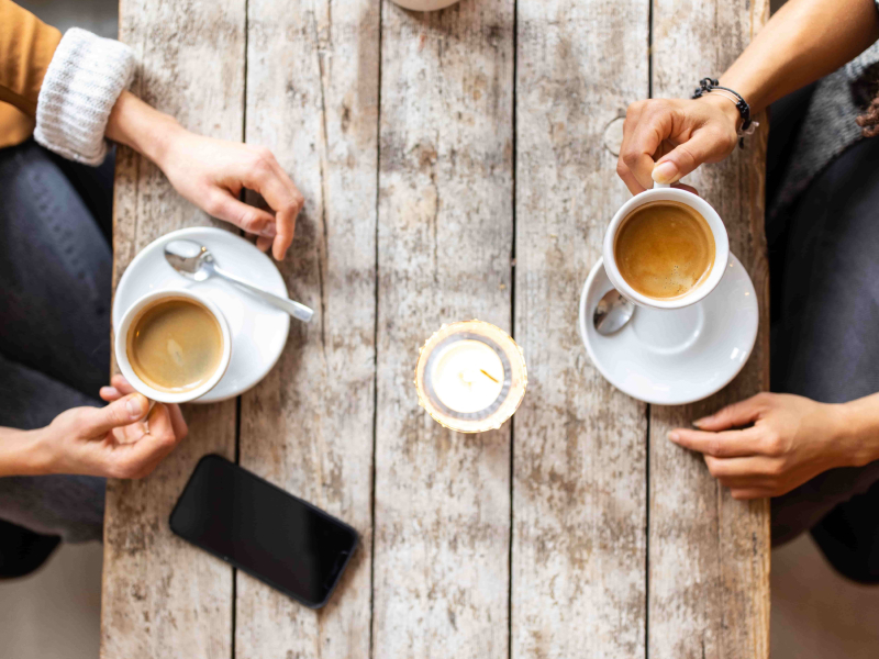 Draufsicht auf einen Tisch, an dem zwei Frauen jeweils einen Kaffee trinken. Auf dem Tisch liegen außerdem eine Kerze und ein Smartphone.