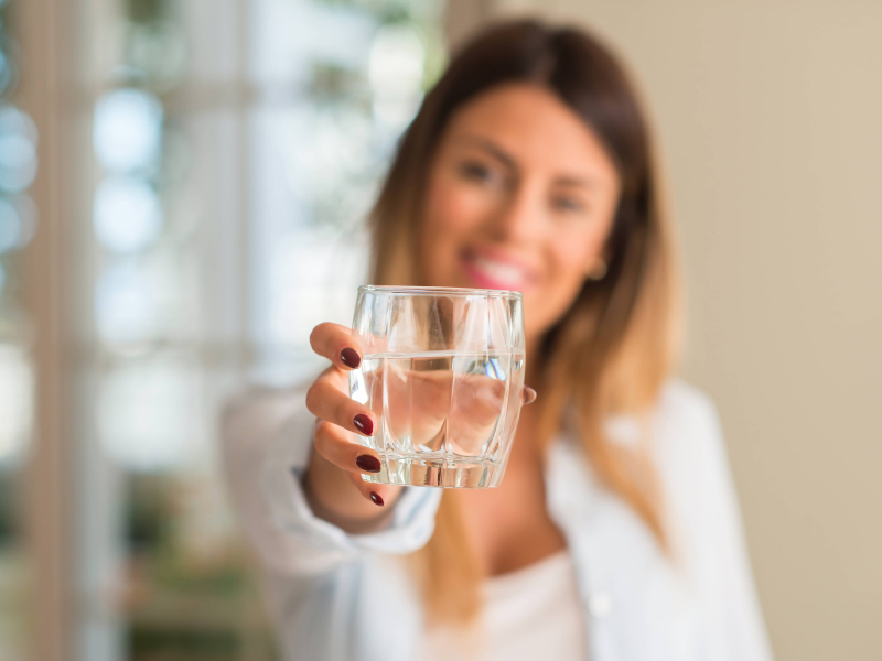 Eine lächelnde Frau hält ein Glas mit Wasser in die Kamera. Das Glas ist fokussiert im Vordergrund, die Frau im Hintergrund unscharf.