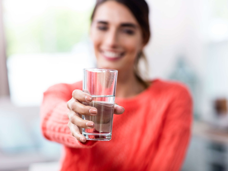 Eine verschwommene Frau im Hintergrund mit rot-pinkem Pulli hält ein Glas Wasser in den Fokus.