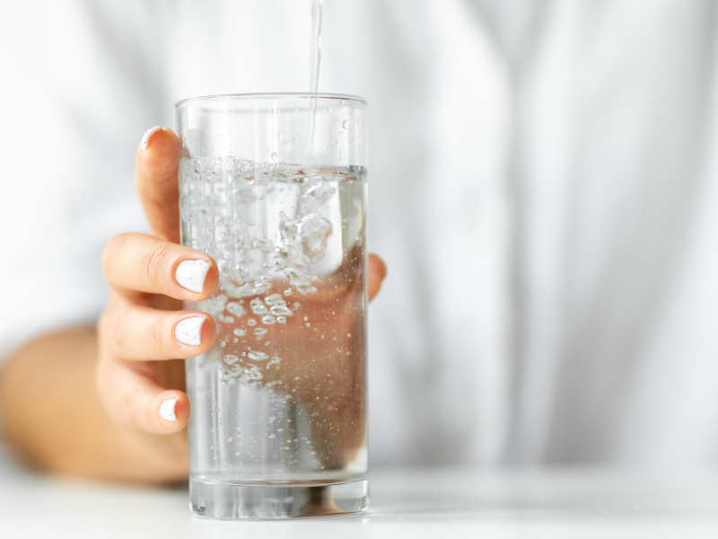 Eine Person mit weißlackierten Fingernägeln hält ein Glas in der Hand, in der gerade Sprudelwasser eingegossen wird.