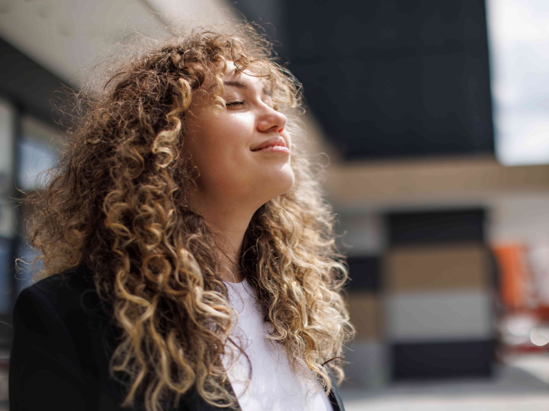 Frau mit Locken steht an der frischen Luft, hält das Gesicht in die Sonne und atmet tief durch.