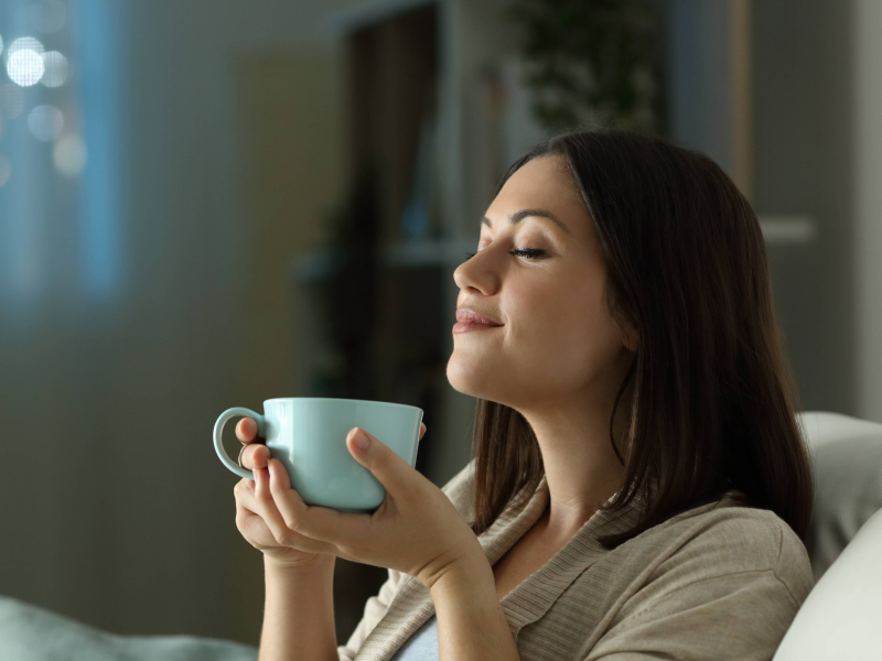 Frau sitzt entspannt mit geschlossenen Augen auf dem Sofa und hält eine Tasse mit einem warmen Getränk in der Hand.