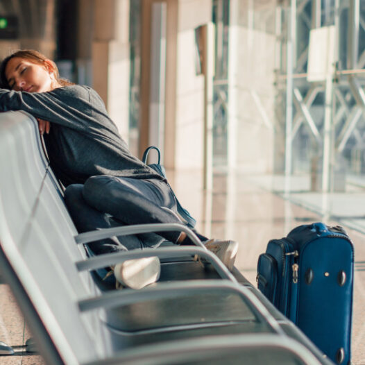 Müde Frau schläft auf Sitzbank am Flughafen. Neben ihr steht ein blauer Koffer.
