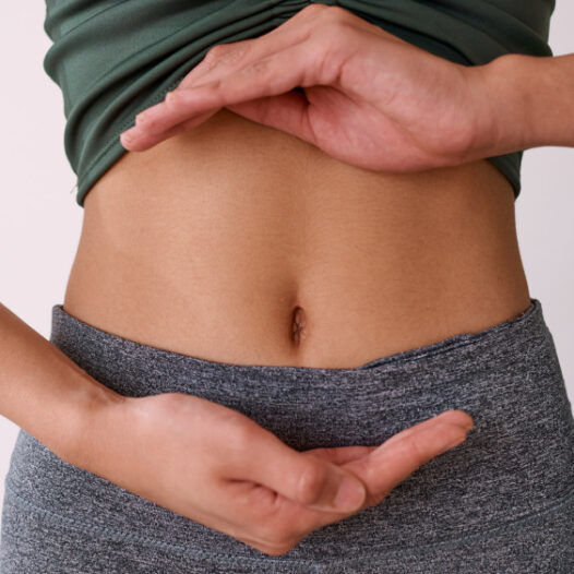 Nahaufnahme auf den Bauch einer jungen Person. Die Hände liegen jeweils oben und unten am Bauch mit den Handflächen zueinander.
