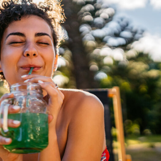 Junge Frau ist bei Sonnenschein draußen und trinkt lächelnd einen grünen Smoothie.