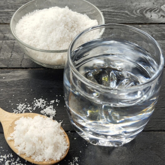 Ein Glas Wasser und eine Schüssel Salz mit einem Holzlöffel voll Salz liegen auf einem Holztisch.
