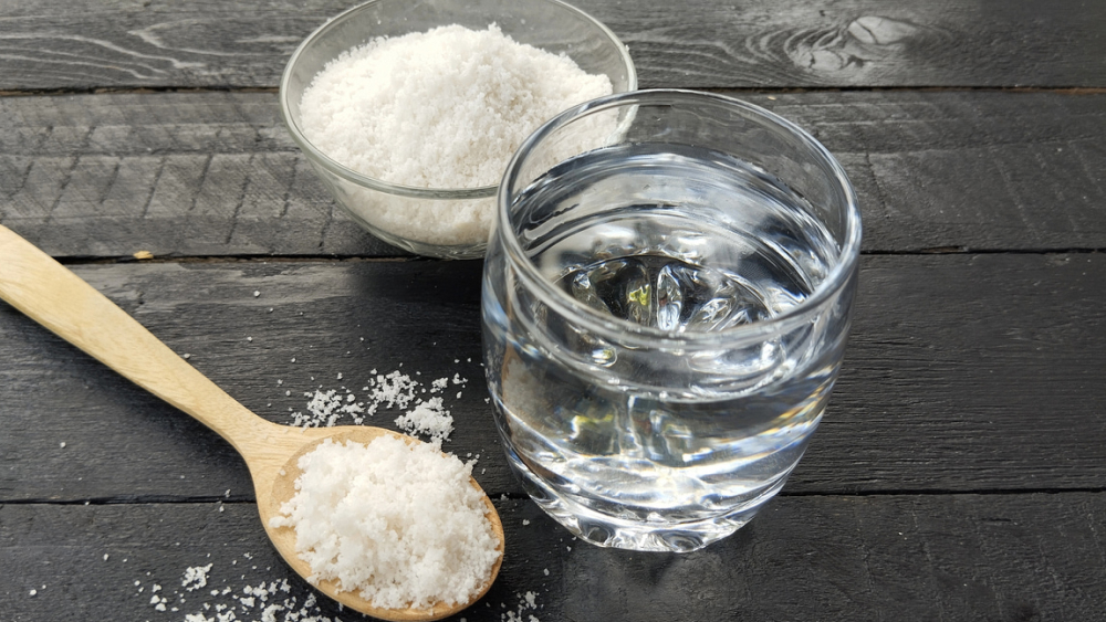 Ein Glas Wasser und eine Schüssel Salz mit einem Holzlöffel voll Salz liegen auf einem Holztisch.