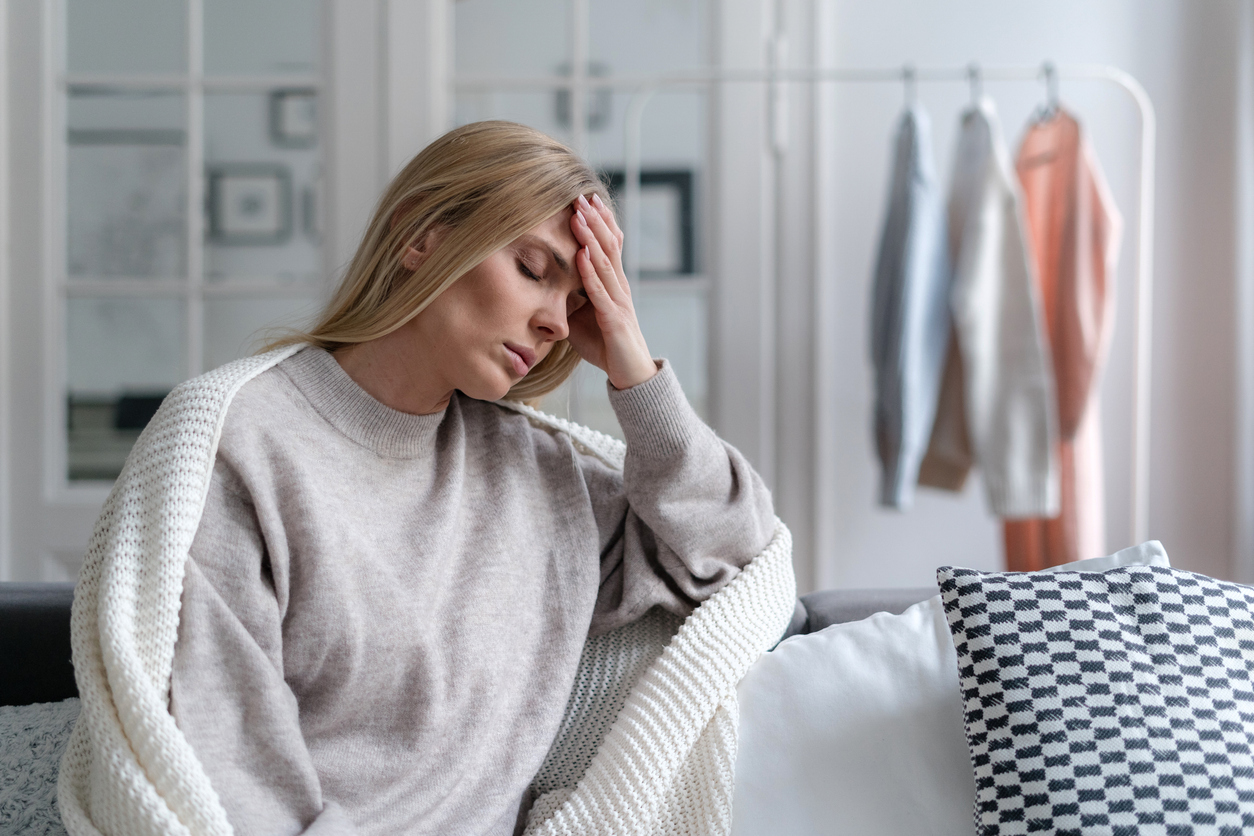 Junge, blonde Frau sitzt zu Hause in eine Decke eingehüllt auf dem Sofa und zeigt Anzeichen von Unwohlsein